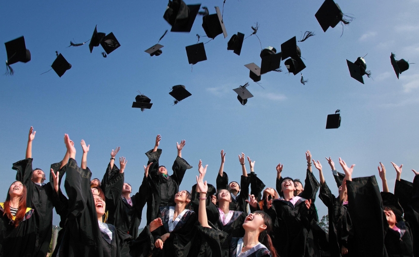 Nhóm sinh viên tốt nghiệp mặc áo thạc sĩ đen, ném mũ tốt nghiệp (tassel) lên trời, tay giơ cao, cười rạng rỡ, dưới bầu trời xanh trong, ánh sáng tự nhiên ban ngày.