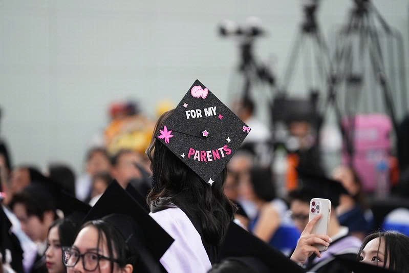 Một sinh viên đội mũ tốt nghiệp (mortarboard) hình vuông, trang trí kim tuyến đen, dán chữ nổi 'FOR MY PARENTS' và họa tiết ngôi sao hồng, trong buổi lễ đông người với ánh sáng hội trường.