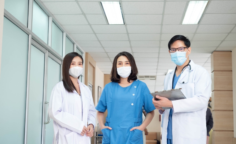 Ba nhân viên y tế (hai nữ, một nam) đứng trong hành lang bệnh viện: nữ bên trái mặc áo blouse trắng, nữ giữa mặc scrubs xanh dương, nam bên phải mặc blouse trắng khoác ngoài scrubs xanh, đeo ống nghe