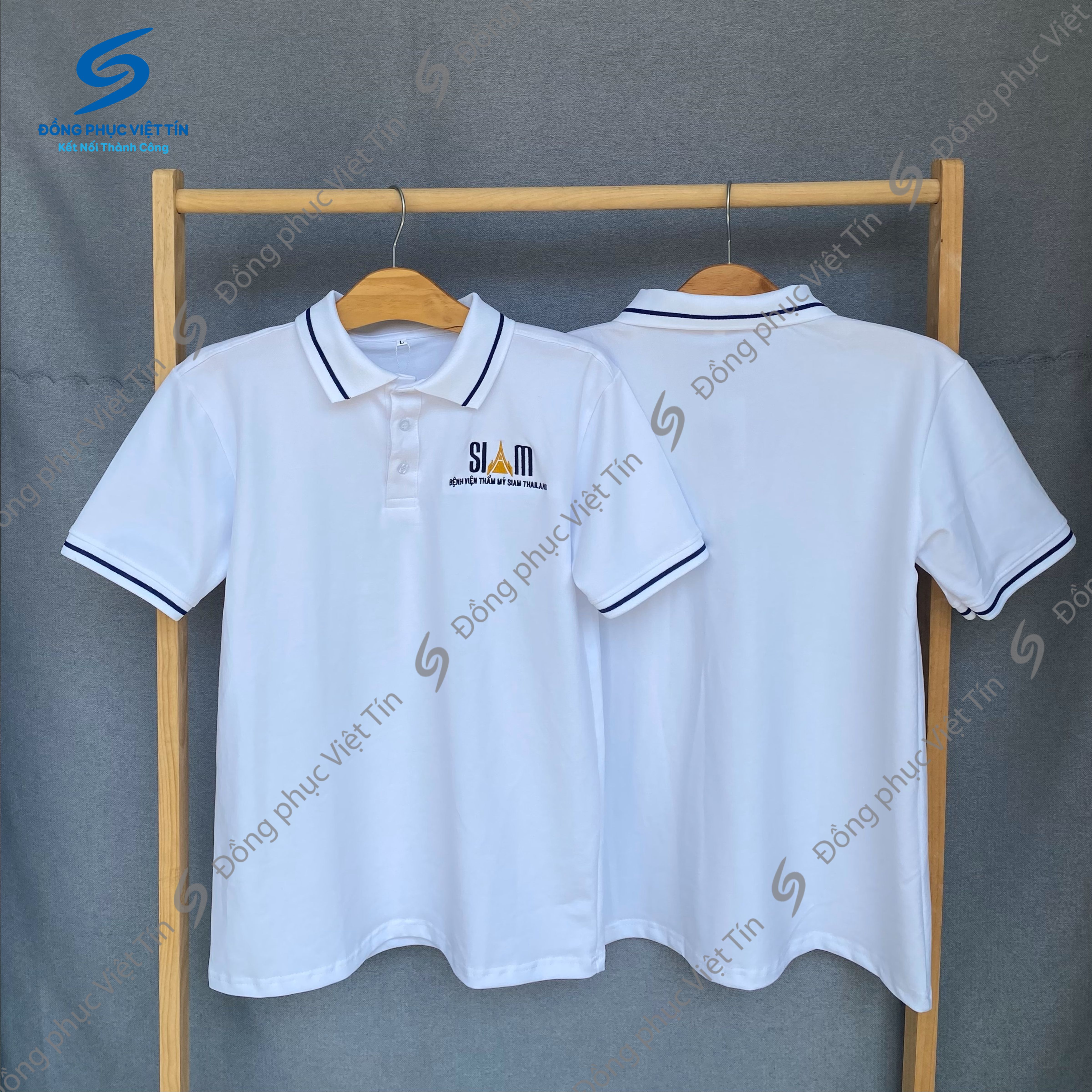 Hai chiếc áo polo màu trắng có cổ và viền tay màu xanh đen, chất liệu Cá sấu Cotton 100%, treo trên giá gỗ. Áo có thêu logo 'SIAM' ở ngực trái. Nền xám, có watermark thương hiệu.