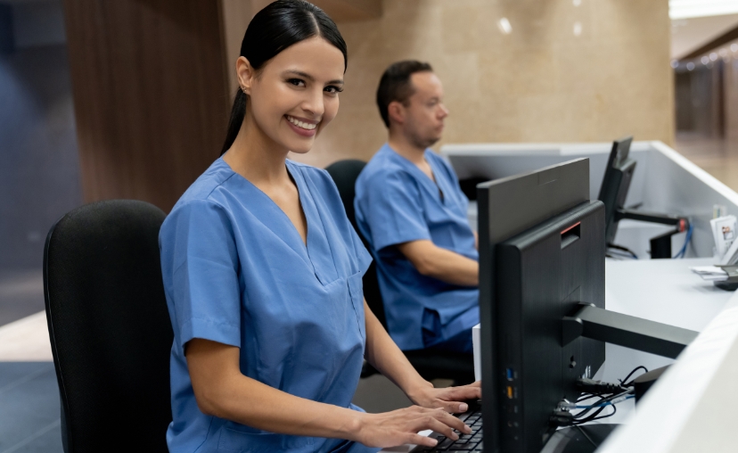 Hai nhân viên y tế mặc đồng phục màu xanh dương ngồi tại bàn làm việc với máy tính. Người phụ nữ ở tiền cảnh mỉm cười nhìn vào ống kính, tay đặt trên bàn phím. Đồng phục có cổ chữ V, tay ngắn, chất li