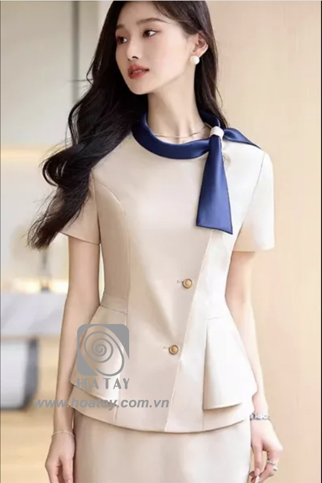 Một nữ nhân viên đang đứng, mặc áo đồng phục công sở màu be (beige) tay ngắn, cổ tròn phối khăn lụa xanh dương, thiết kế vạt chéo (asymmetrical) với hai cúc kim loại, dáng peplum xòe eo, ánh sáng tự n