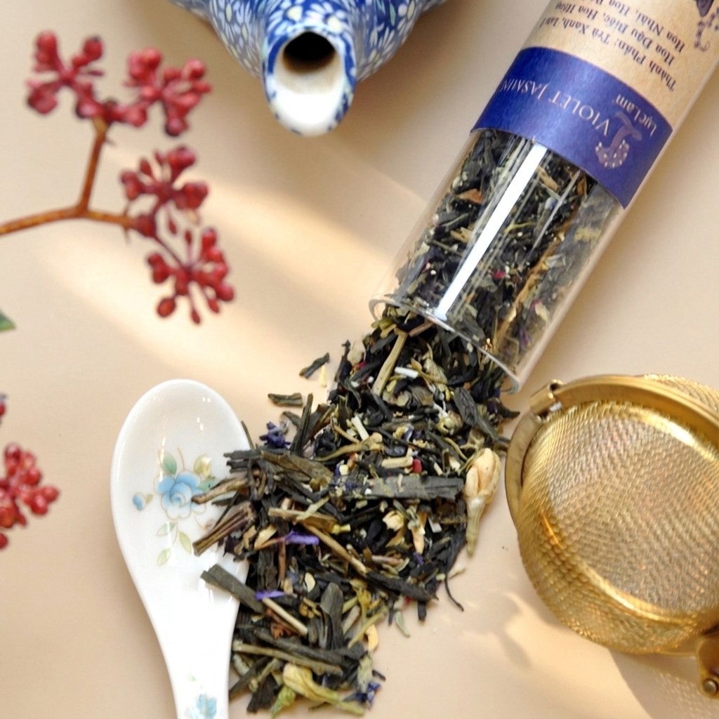 Loose leaf tea blend (1) spilling from a clear cylindrical glass tube: dried green tea leaves, jasmine buds, and purple flower petals, accompanied by a gold mesh tea infuser and a small ceramic spoon.