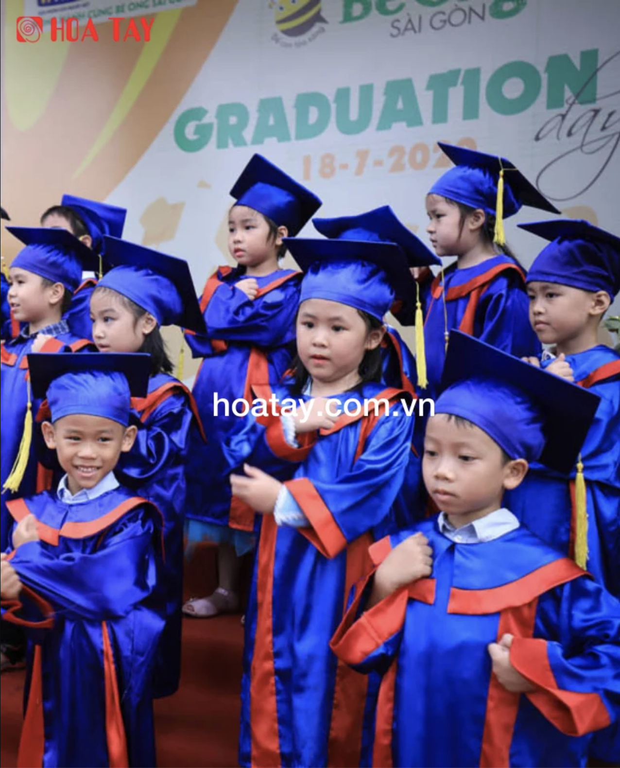 Nhóm trẻ em (10+) đang đứng nghiêm trang, mặc áo cử nhân (áo tốt nghiệp) màu xanh dương phối viền đỏ, mũ vuông (nón tốt nghiệp) có tua rua vàng, chất liệu vải bóng, bối cảnh lễ tốt nghiệp mầm non, ánh