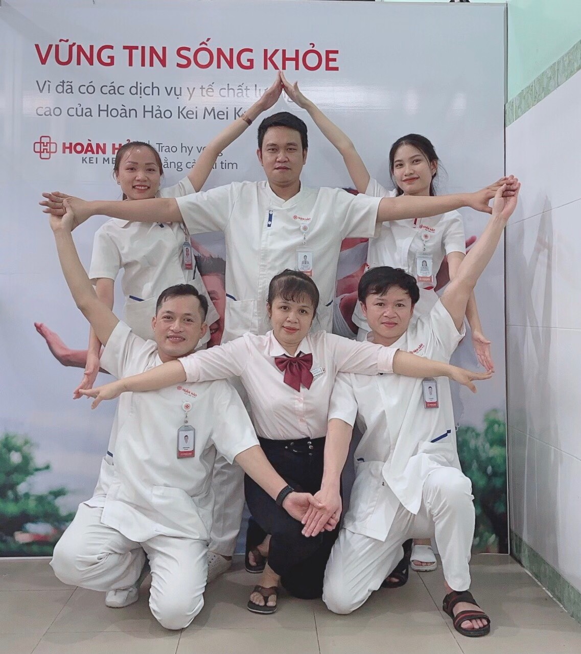 Sáu nhân viên y tế (năm người mặc áo blouse trắng, một người mặc sơ mi hồng) đang tạo dáng hình ngôi nhà bằng tay; áo blouse cổ bẻ, tay ngắn, có bảng tên, đứng trước phông nền quảng cáo bệnh viện, ánh
