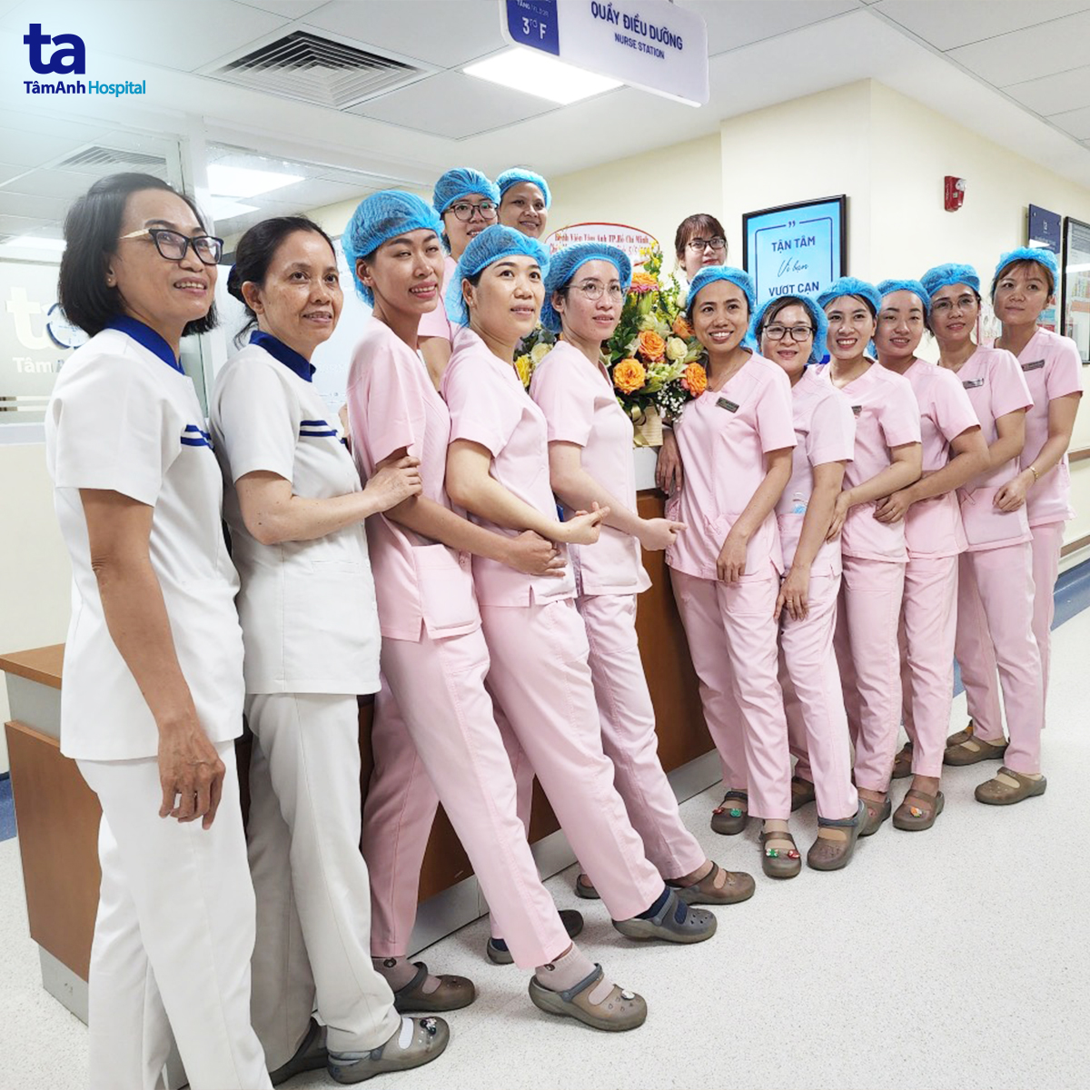 Nhóm 12 nữ điều dưỡng (y tá) đứng thành hàng ngang tại quầy trực; mặc đồng phục hồng và trắng, đội mũ bao tóc (nón y tế) màu xanh, áo cổ chữ V, quần ống đứng; ánh sáng đèn huỳnh quang trong bệnh viện.