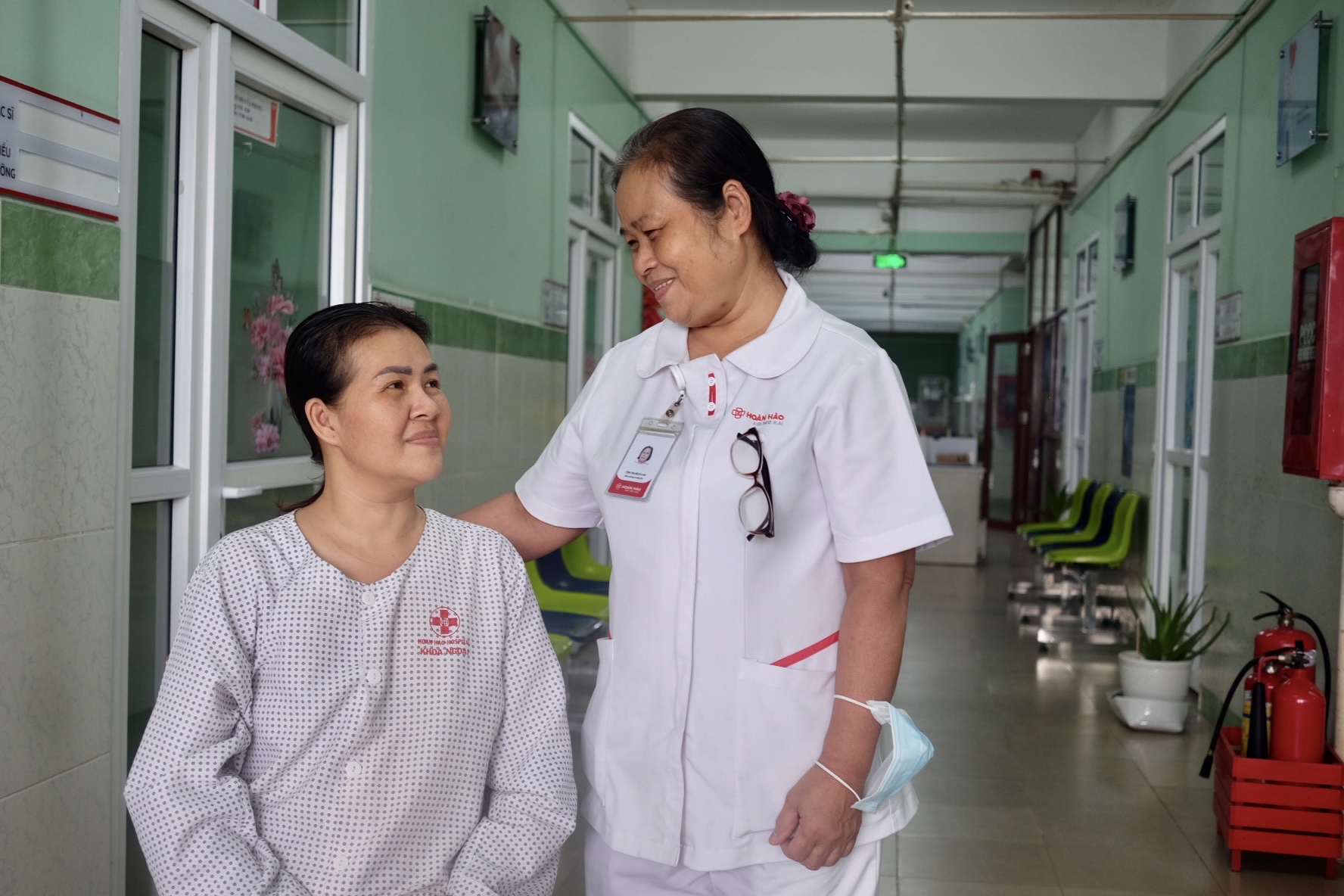 Một nữ y tá và một bệnh nhân nữ đang đứng trò chuyện. Y tá mặc áo blouse trắng cổ bẻ (cổ lá sen), cài thẻ tên, đeo kính. Bệnh nhân mặc đồ bệnh viện họa tiết chấm bi nhỏ. Hành lang bệnh viện sáng đèn,