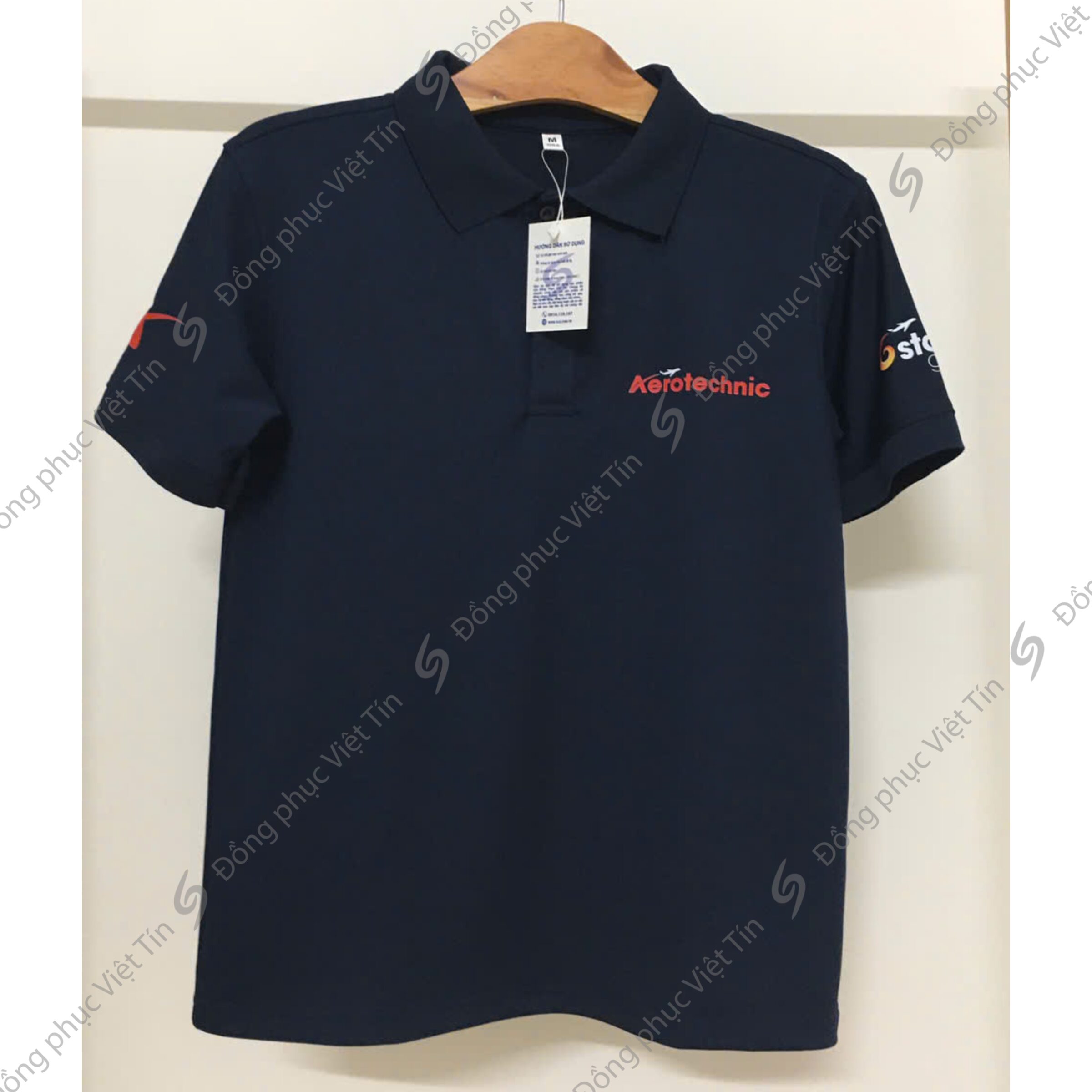 Áo polo màu xanh đen cổ bẻ, treo trên móc gỗ. Áo có logo Aerotechnic màu đỏ ở ngực trái và logo thương hiệu ở tay áo. Hình ảnh có watermark chéo của Đồng phục Việt Tín.