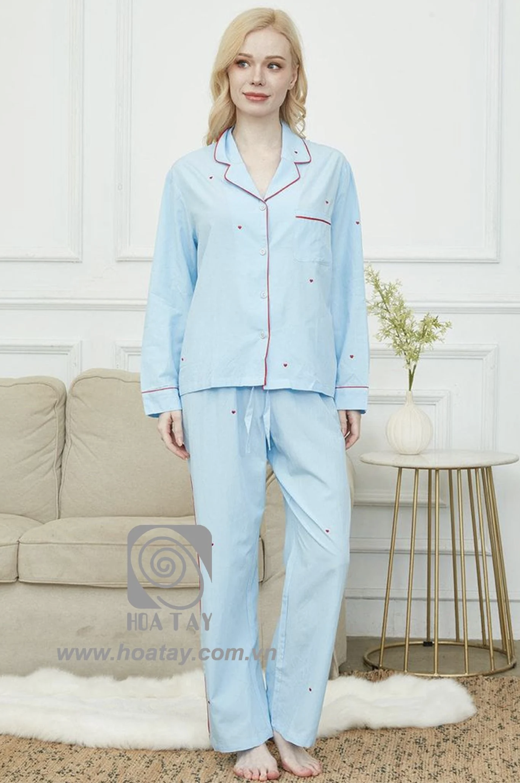 Một phụ nữ mặc bộ đồ ngủ (pajamas) màu xanh nhạt, họa tiết trái tim đỏ nhỏ, cổ bẻ (notch lapel), cài cúc phía trước, chất liệu cotton mềm, đứng trong phòng khách với ánh sáng tự nhiên.