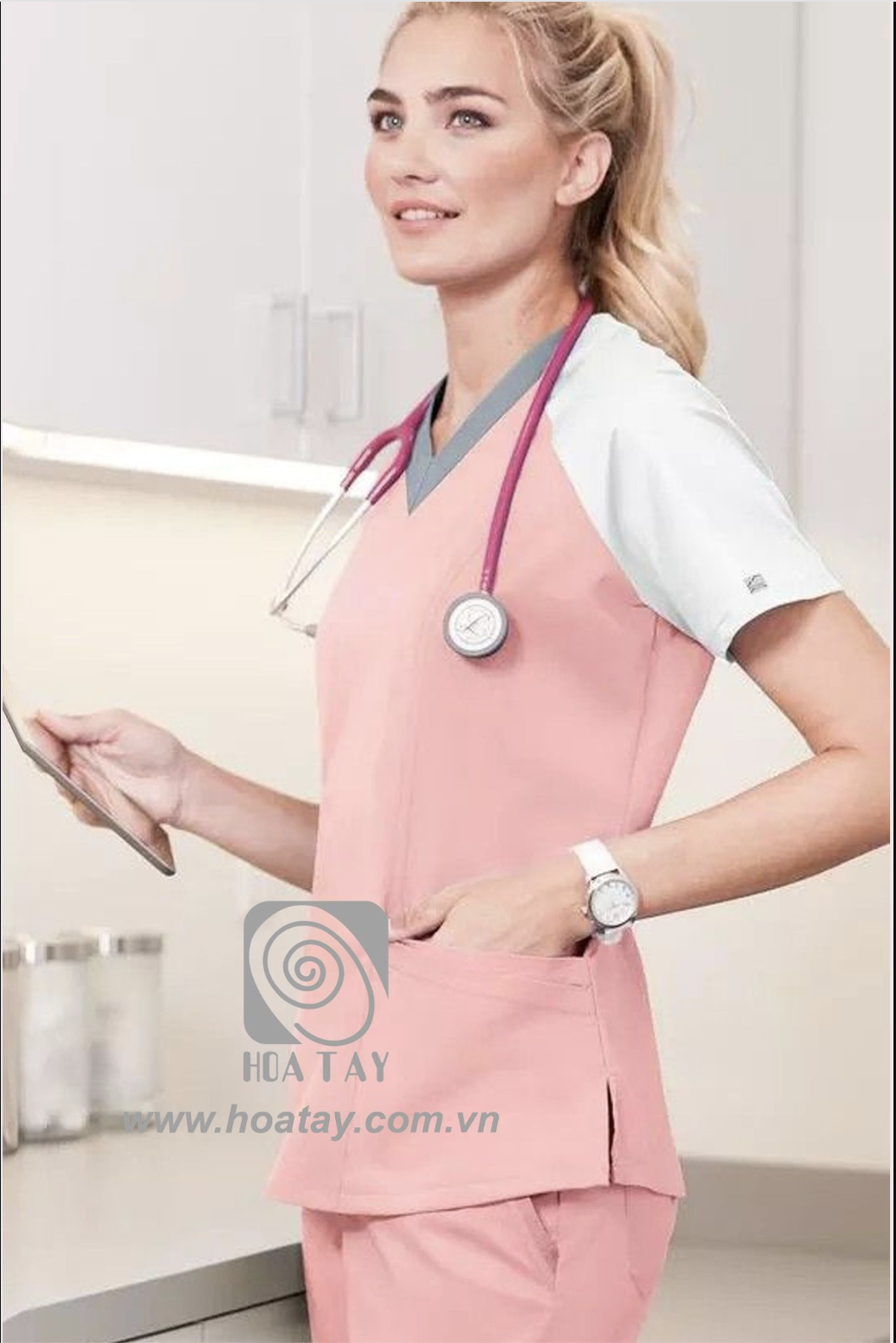 Một nữ nhân viên y tế đang đứng; mặc áo cổ chữ V (v-neck) phối màu hồng-trắng, chất liệu vải mềm, có túi hông, ống tay ngắn, đeo ống nghe y tế; bối cảnh phòng khám sáng sủa.