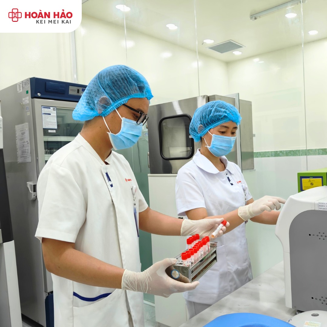 Hai kỹ thuật viên xét nghiệm (hai nhân viên) đang vận hành máy phân tích, mặc áo blouse trắng (áo khoác phòng thí nghiệm), đội mũ bao tóc xanh, đeo khẩu trang y tế, găng tay cao su, trong phòng lab sá