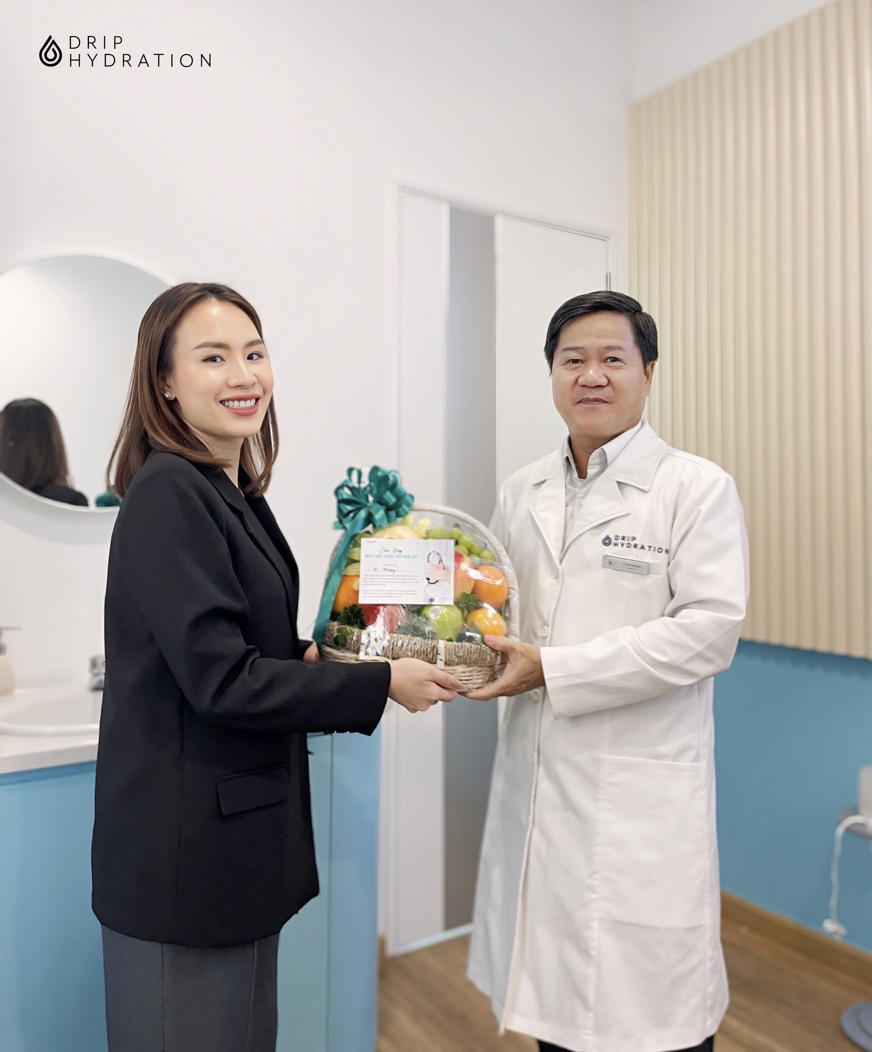 Hai người (một nam, một nữ) đang trao giỏ quà trái cây. Người nam mặc áo blouse trắng (áo khoác y tế) có logo, người nữ mặc blazer đen. Bối cảnh phòng khám sạch sẽ, ánh sáng tự nhiên.