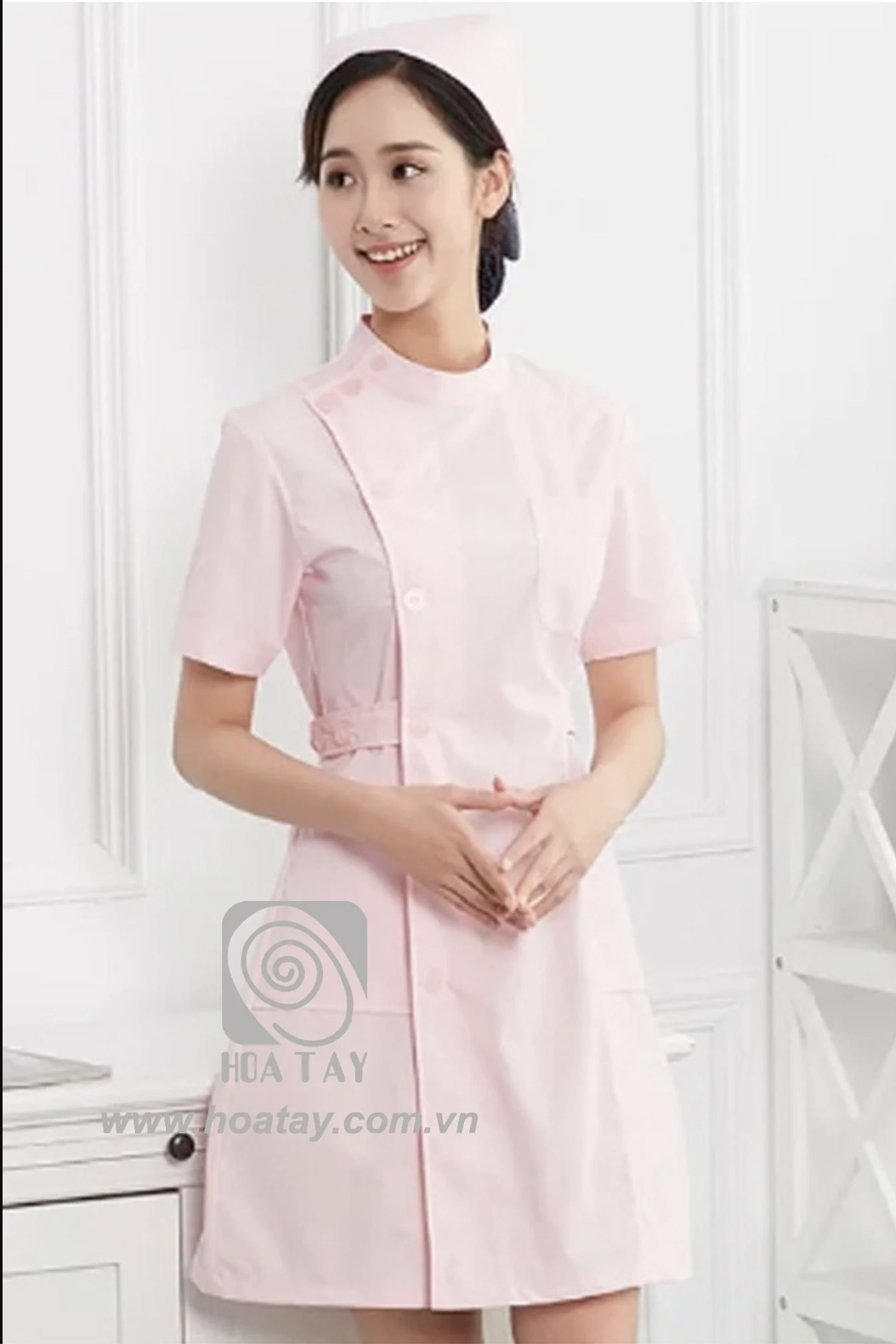 Một nữ điều dưỡng đang đứng, mặc áo blouse hồng nhạt, cổ tàu (cổ đứng), thiết kế vạt chéo (cài cúc lệch) tay ngắn, chất vải cotton phẳng phiu, đội mũ y tế cùng tông màu, bối cảnh studio ánh sáng trắng