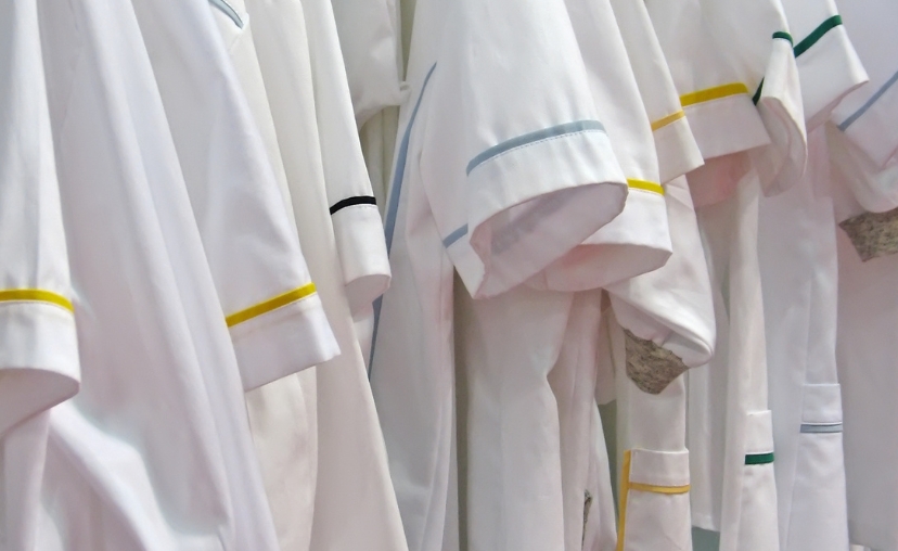 Nhiều áo blouse trắng treo trên giá, tay áo ngắn với viền piping màu vàng, xanh dương và xanh lá, chất liệu vải trắng trơn, ánh sáng tự nhiên.
