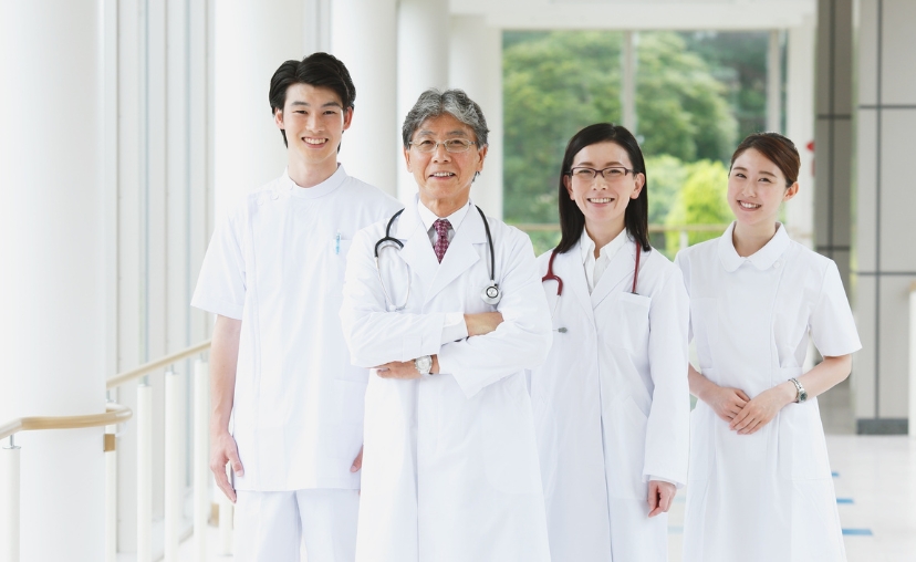 Bốn nhân viên y tế (hai nam, hai nữ) đứng hàng ngang trong hành lang bệnh viện. Người đàn ông lớn tuổi mặc áo blouse trắng dài, đeo ống nghe, tay khoanh. Ba người còn lại mặc áo blouse trắng ngắn tay.