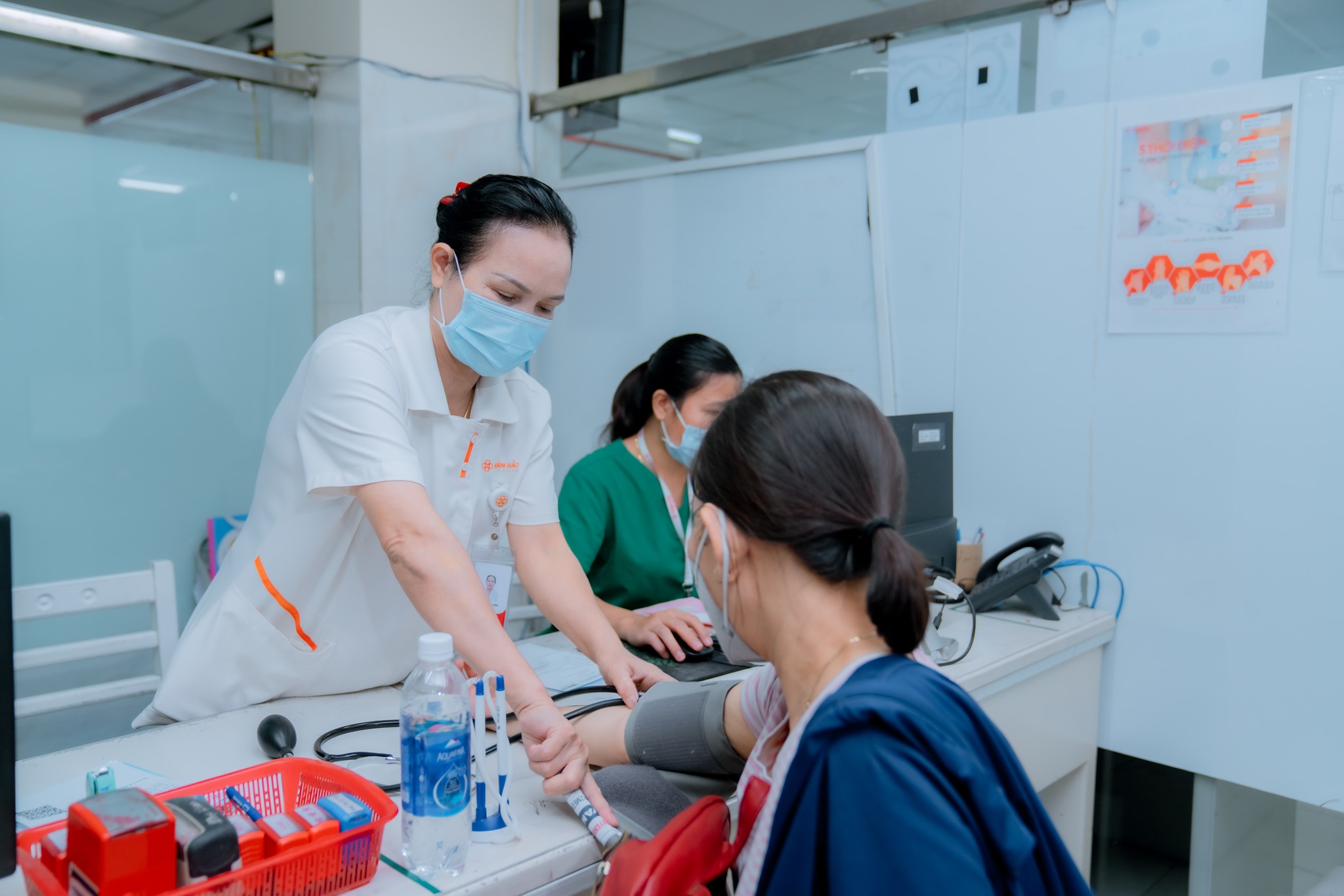 Một nữ nhân viên y tế đang đo huyết áp cho bệnh nhân; mặc áo blouse trắng (áo khoác y tế), đeo khẩu trang y tế, có thẻ tên, chi tiết viền cam, chất liệu vải thô, bối cảnh phòng khám bệnh viện, ánh sán