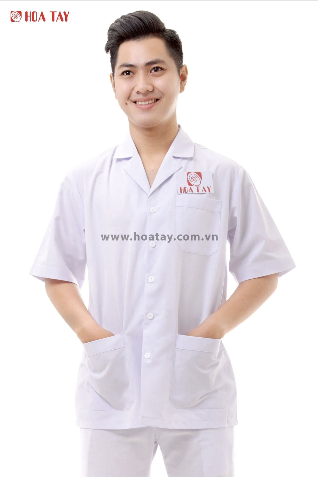 Một nam nhân viên đang đứng cười, mặc áo blouse trắng (áo bác sĩ) tay ngắn, cổ bẻ (cổ vest), chất liệu vải cotton phẳng, có túi ngực và hai túi hông, logo thêu đỏ trên nền trắng, phông nền studio sáng
