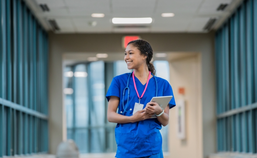 Một nữ y tá -> đi bộ và mỉm cười nhìn sang bên -> mặc bộ đồng phục (scrubs) màu xanh dương, ống nghe màu xanh, dây đeo thẻ màu hồng, cầm máy tính bảng -> hành lang bệnh viện với ánh sáng trắng và cửa
