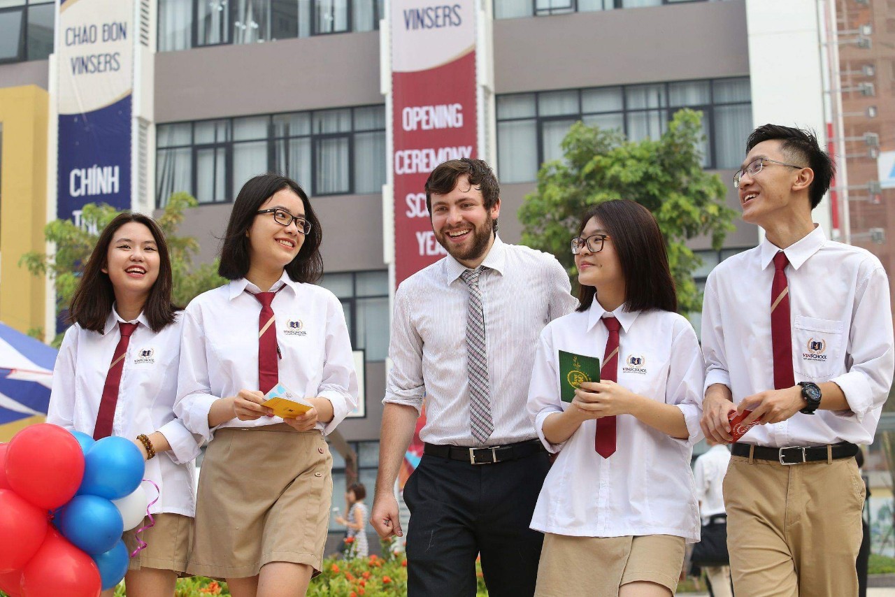 5 người (4 sinh viên, 1 giáo viên) đi bộ ngoài trời. Sinh viên mặc áo sơ mi trắng, cà vạt đỏ, quần/áo dài màu be, đeo huy hiệu trường. Giáo viên mặc áo sọc, cà vạt kẻ. Nền có cờ hiệu 'VINSERS' và 'OPE