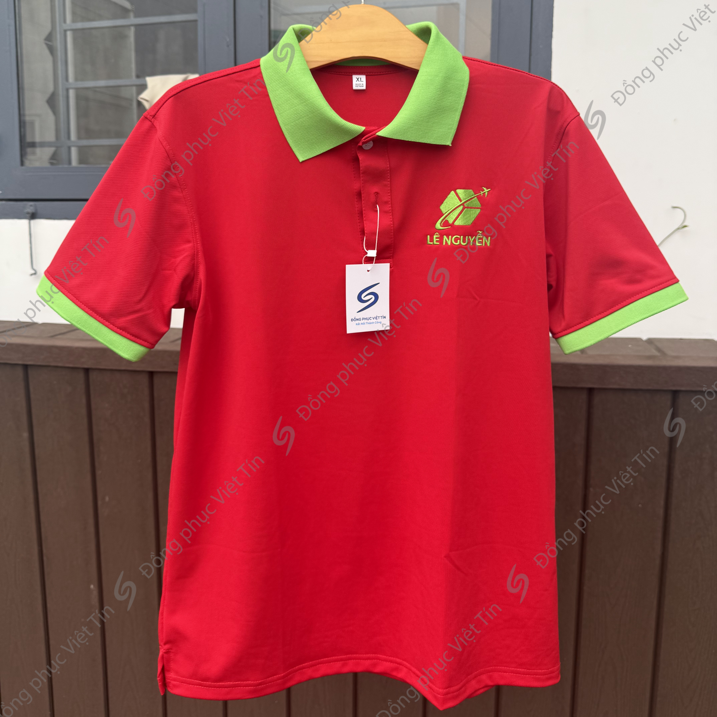 Áo polo màu đỏ với cổ và viền tay áo màu xanh lá, chất liệu Poly cao cấp, có logo thêu 'LÊ NGUYỄN' phía trước, treo trên móc gỗ, nền tường gỗ nâu.