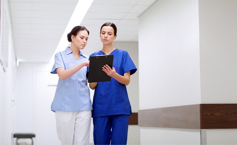 Hai nữ y tá đứng trong hành lang bệnh viện, cùng nhìn vào một bảng kẹp (clipboard) đen. Một người mặc áo sơ mi cổ bẻ màu xanh nhạt và quần trắng; người kia mặc bộ đồng phục (scrubs) màu xanh cobalt vớ
