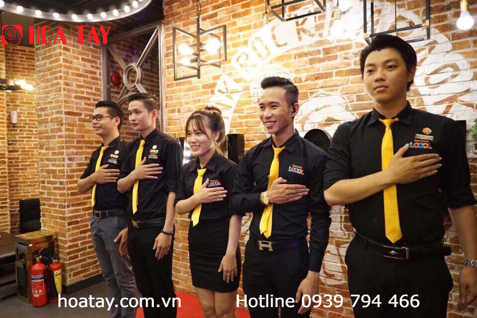 Năm nhân viên (ba nam, hai nữ) đang đứng nghiêm, đặt tay lên ngực. Họ mặc sơ mi đen dài tay, cà vạt vàng, quần tây hoặc chân váy đen. Bối cảnh tường gạch thô, ánh sáng vàng ấm áp từ đèn treo.