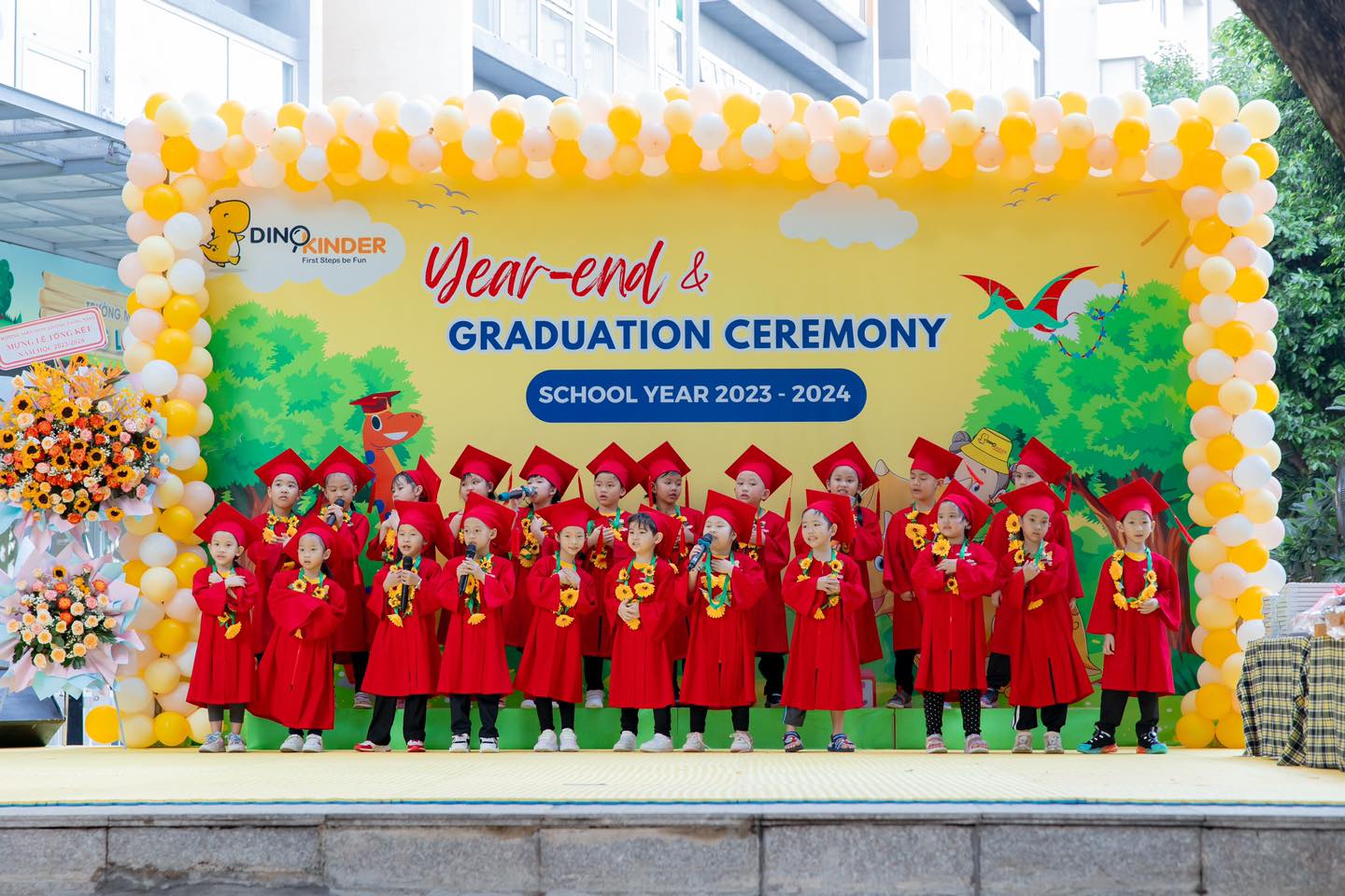 Nhóm 20 trẻ mầm non đang đứng hát trên sân khấu; mặc áo cử nhân (áo tốt nghiệp) màu đỏ, mũ vuông, vòng hoa hướng dương; phông nền trang trí bóng bay vàng trắng và bảng hiệu Year-end & Graduation Cerem