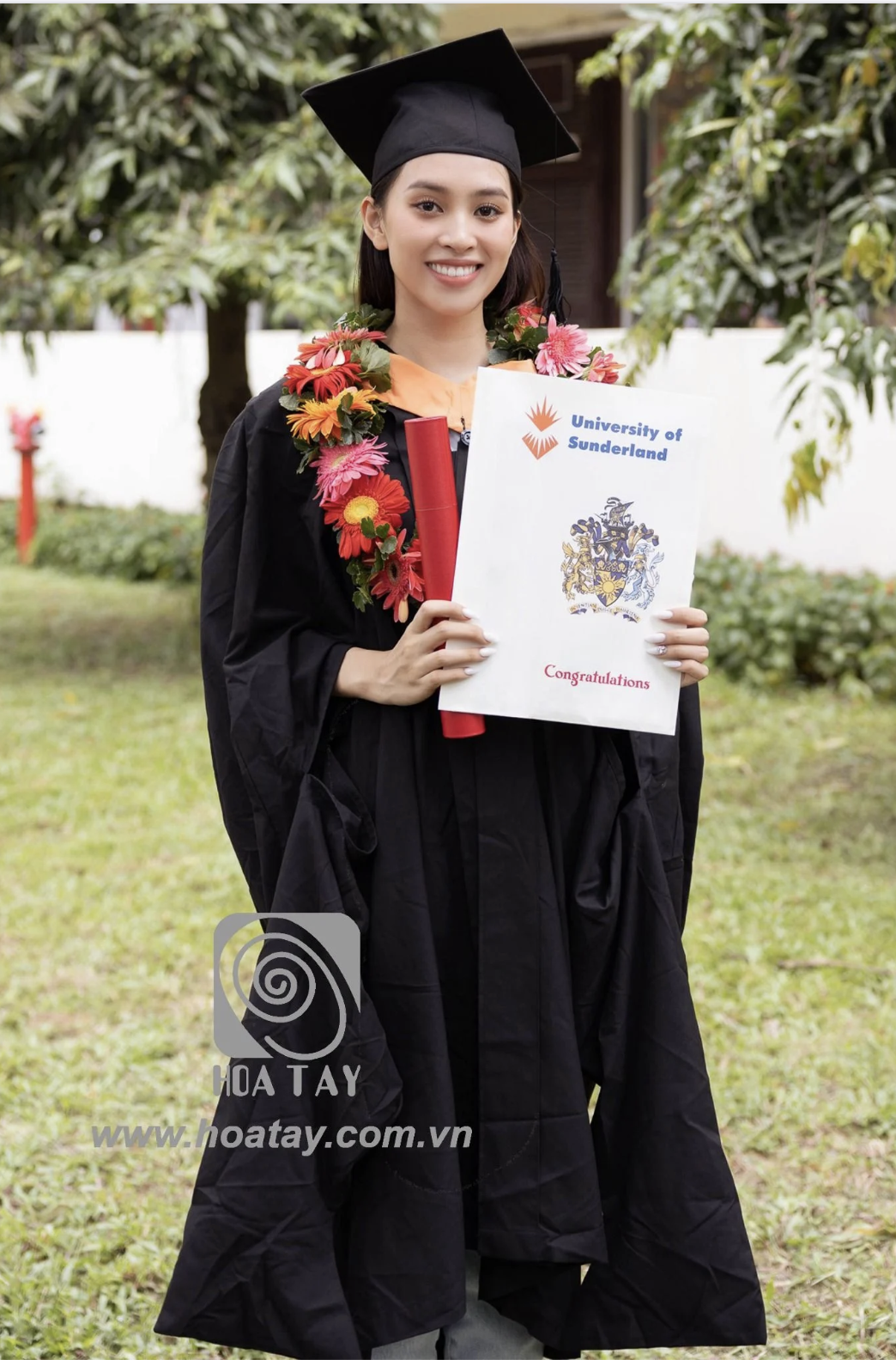 Một nữ sinh viên tốt nghiệp đang đứng cười; mặc áo cử nhân (áo thụng) đen bằng vải polyester, đội mũ mortarboard (mũ vuông) có tua rua, đeo vòng hoa tươi nhiều màu, cầm bằng tốt nghiệp University of S