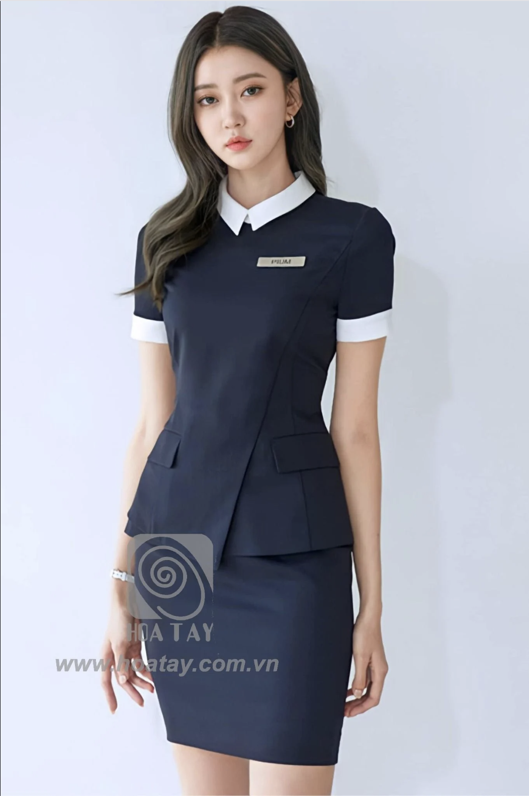 Một nữ nhân viên đứng thẳng, mặc đồng phục váy liền (dress) màu xanh đen, cổ áo sơ mi trắng, tay áo ngắn viền trắng, thiết kế vạt chéo (asymmetrical wrap) với hai túi giả, chất liệu vải phẳng mịn, phô