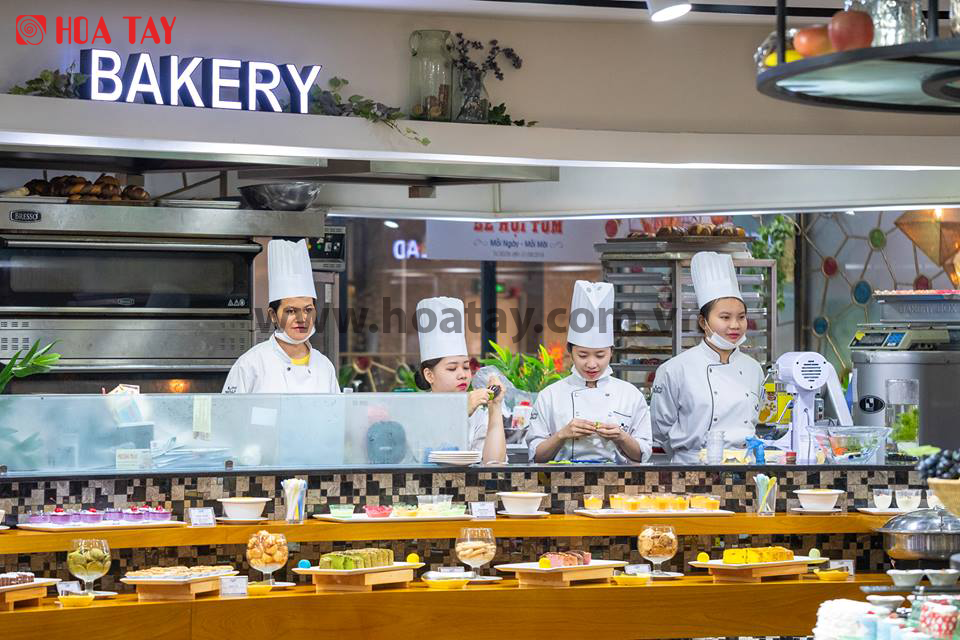 Bốn thợ làm bánh (đầu bếp) đang làm việc trong bếp mở; mặc áo khoác trắng cài khuy chéo (double-breasted), mũ cao (toque), khẩu trang y tế; không gian tiệm bánh hiện đại, ánh sáng đèn huỳnh quang rực
