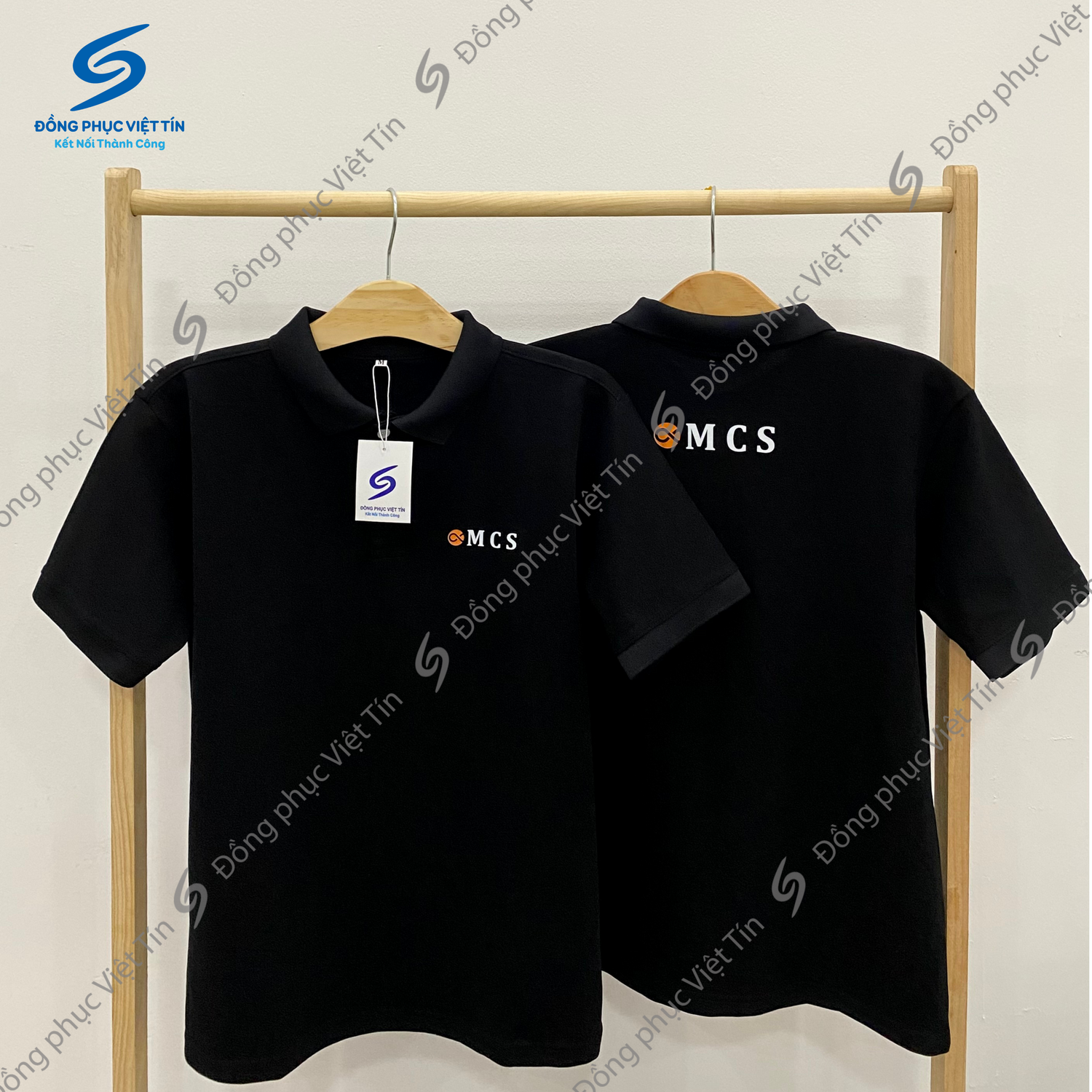 Hai chiếc áo polo màu đen có in logo MCS, treo trên giá gỗ, nền trắng. Áo có cổ, tay ngắn, chất liệu Cá sấu Cotton 100%, dày dặn, đứng form, thấm hút mồ hôi tốt.
