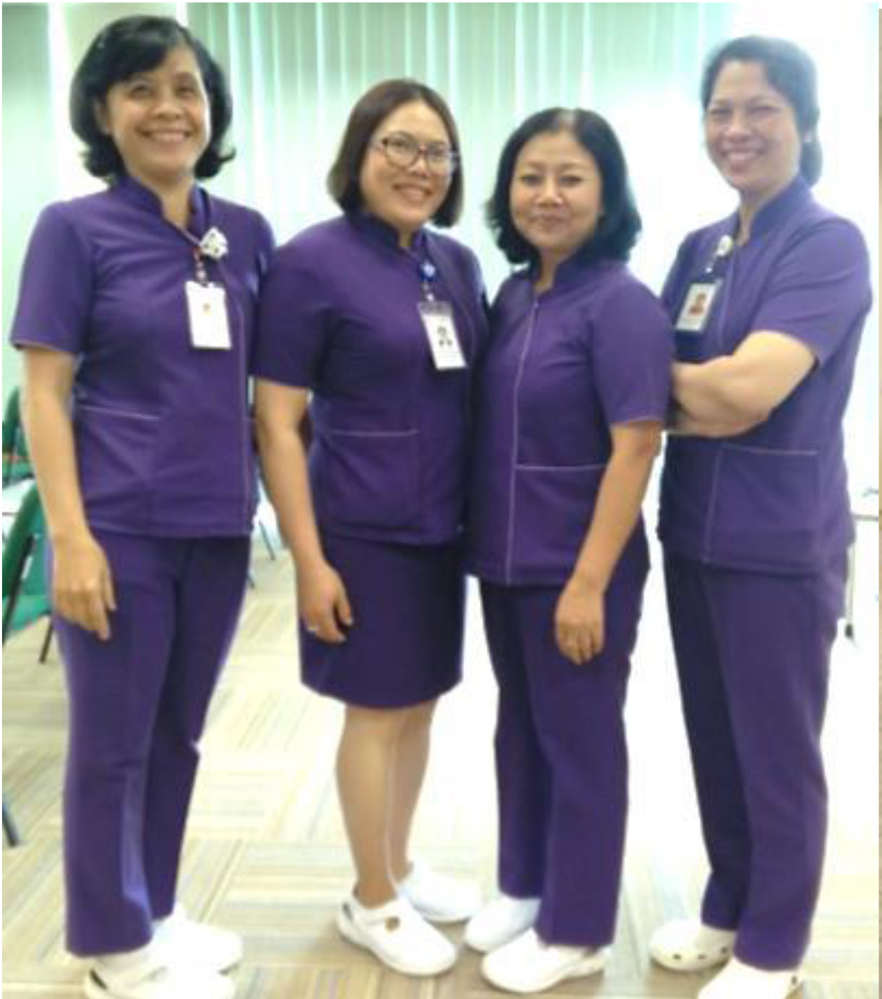 Bốn nữ nhân viên y tế đứng cạnh nhau, mặc đồng phục y tá màu tím gồm áo cổ đứng và quần dài hoặc váy, đeo thẻ nhân viên, đi giày trắng, trong phòng có sàn gỗ và rèm sáng.