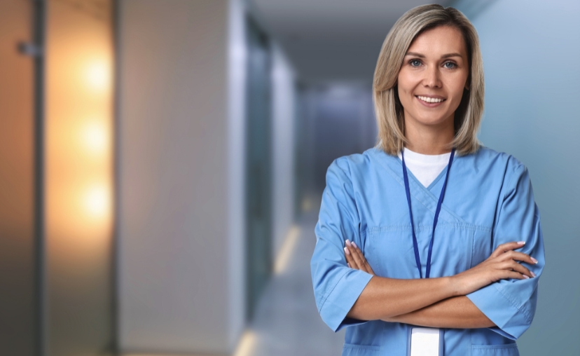 Một nữ y tá tóc vàng, đứng với hai tay khoanh trước ngực, mặc bộ đồng phục y tế (scrubs) màu xanh dương, cổ chữ V, tay lửng, đeo dây đeo thẻ màu xanh đậm, đứng trong hành lang bệnh viện với ánh sáng ấ