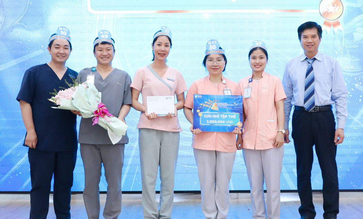 Sáu nhân viên y tế (năm nữ, một nam) đứng hàng ngang, đang nhận giải thưởng; mặc đồng phục scrubs (áo cổ chữ V) màu xanh navy, xám, hồng, đeo thẻ tên, đội mũ giấy in số, cầm hoa và bảng vinh danh; phô