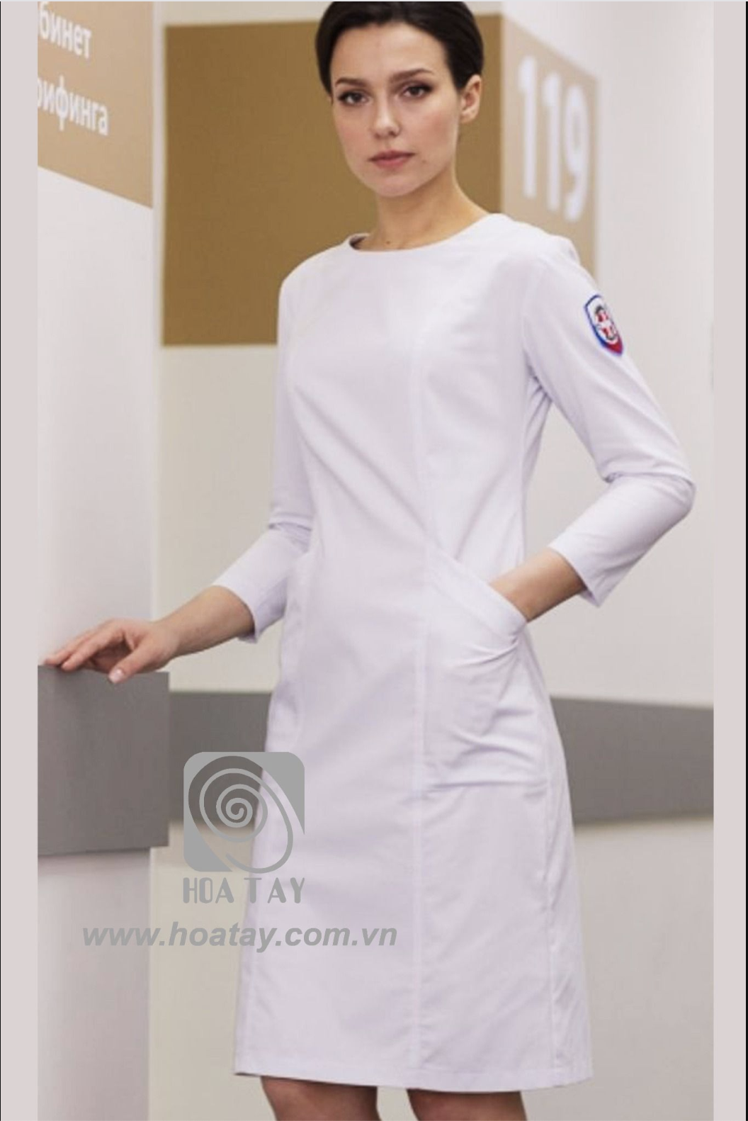 Một nữ nhân viên y tế đứng, mặc áo blouse trắng (áo khoác bác sĩ) dài tay, cổ tròn, chất liệu vải trơn, có hai túi hông chìm, huy hiệu trên tay áo, bối cảnh hành lang bệnh viện với ánh sáng dịu.