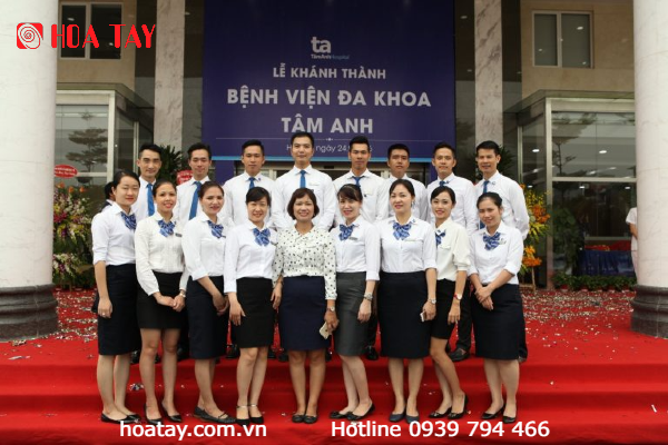 Nhóm nhân viên y tế đứng trên thảm đỏ trước biển hiệu bệnh viện, mặc áo sơ mi trắng, váy đen và cà vạt xanh, tạo dáng chụp ảnh tập thể.