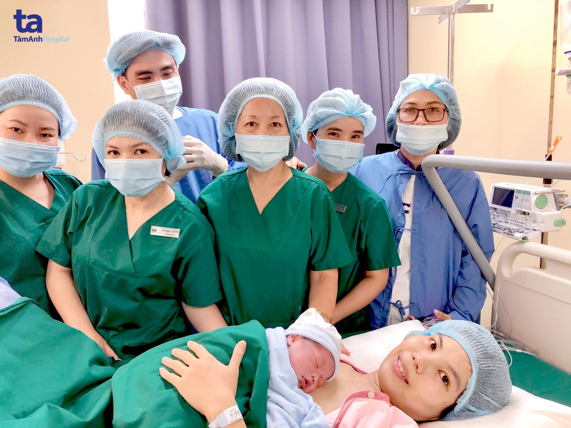 Bảy nhân viên y tế và một sản phụ nằm trên giường bệnh, ôm đứa trẻ sơ sinh. Họ mặc áo phẫu thuật màu xanh lá cây và xanh dương, đội mũ phẫu thuật và đeo khẩu trang y tế. Một người mặc áo choàng chống