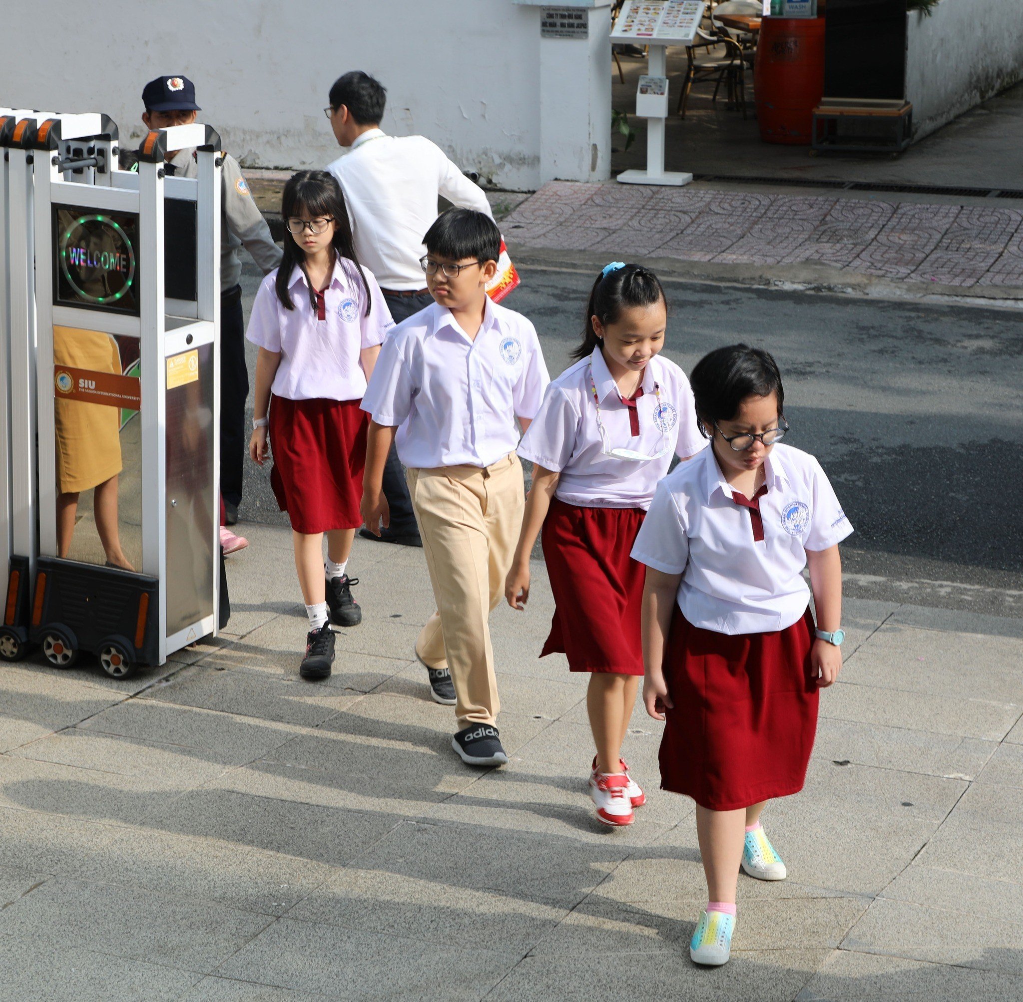 Bốn học sinh (hai nam, hai nữ) đang đi bộ; mặc áo sơ mi trắng ngắn tay (cổ bẻ) và váy/quần màu đỏ hoặc be; đeo kính, đi giày thể thao; bối cảnh sân trường ngoài trời dưới ánh sáng ban ngày.