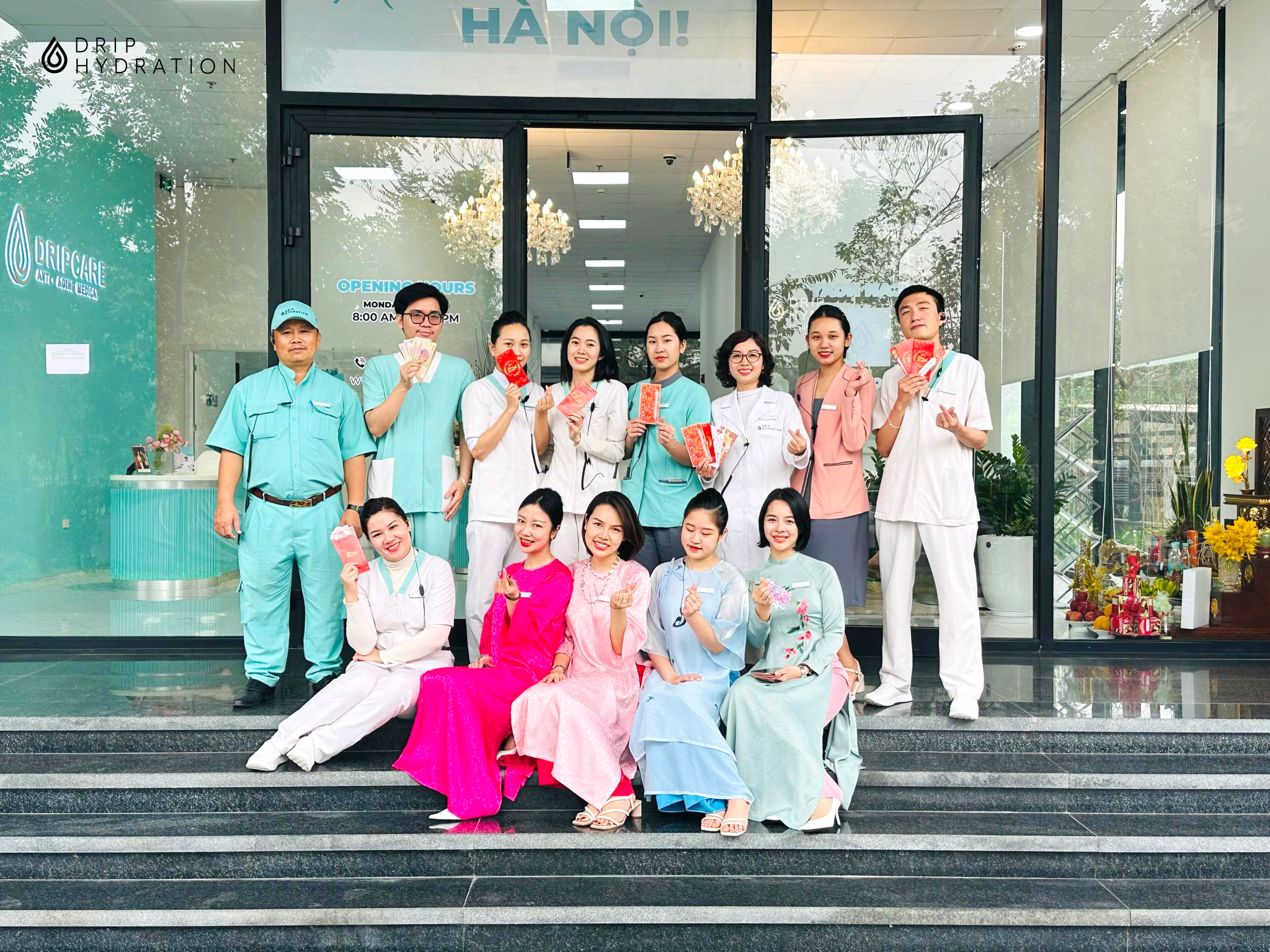 Nhóm 14 nhân viên y tế đang đứng và ngồi trước cửa phòng khám; mặc đồng phục áo blouse trắng, áo scrubs xanh, áo dài truyền thống nhiều màu; bối cảnh tiền sảnh kính hiện đại, ánh sáng tự nhiên ban ngà