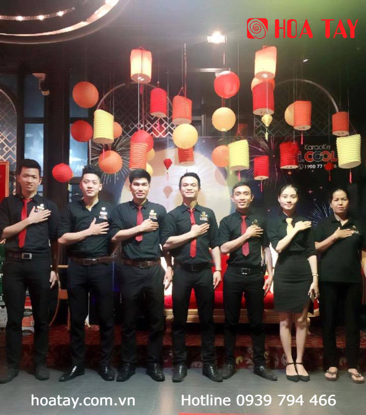 Nhóm 7 nhân viên (đội ngũ) đang đứng đặt tay lên ngực, mặc đồng phục sơ mi đen dài tay (áo sơ mi), cà vạt đỏ, quần tây đen, váy công sở, bối cảnh sự kiện trong nhà với đèn lồng treo trang trí, ánh sán