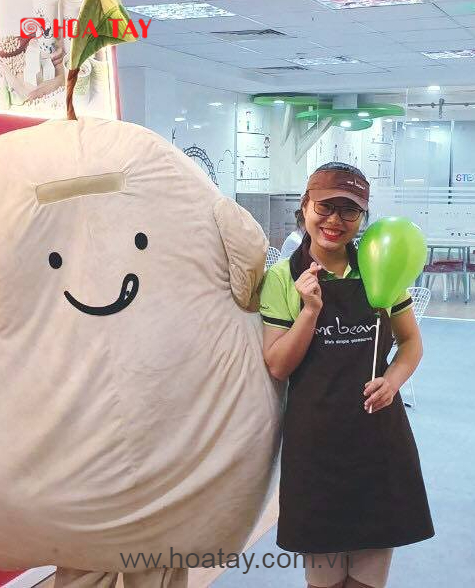 Một nhân viên nữ mặc tạp dề đen (yếm bếp) đứng cạnh một linh vật (mascot) hình hạt đậu màu be, đội mũ lưỡi trai nâu, tay cầm bóng bay xanh lá, bối cảnh cửa hàng sáng đèn.