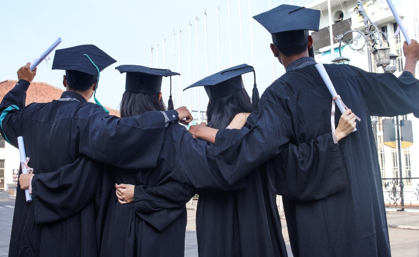 Bốn sinh viên tốt nghiệp quay lưng về phía máy ảnh, mặc áo toga đen và mũ cử nhân vuông có tua rua. Họ ôm vai nhau, tay cầm cuộn bằng trắng. Nền là sân trường ngoài trời với cột cờ và đèn đường, ánh s