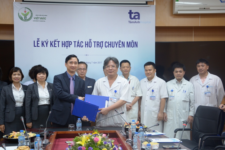 Nhóm 9 người đứng trước phông nền có logo bệnh viện, hai người ở giữa trao nhau một cặp hồ sơ màu xanh dương. Người mặc áo vest, người mặc áo blouse trắng có tên thẻ. Bàn họp phía trước có hoa, nước u