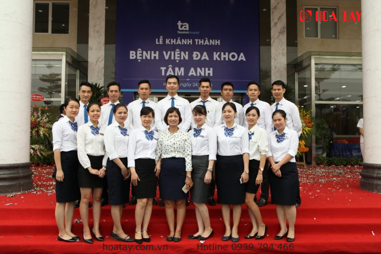Nhóm 15 nhân viên y tế đứng thành hai hàng trên thảm đỏ, mặc áo sơ mi trắng, cà vạt xanh, chân váy đen, đứng trước biển hiệu 'Lễ khánh thành Bệnh viện đa khoa Tâm Anh' với ánh sáng ban ngày.