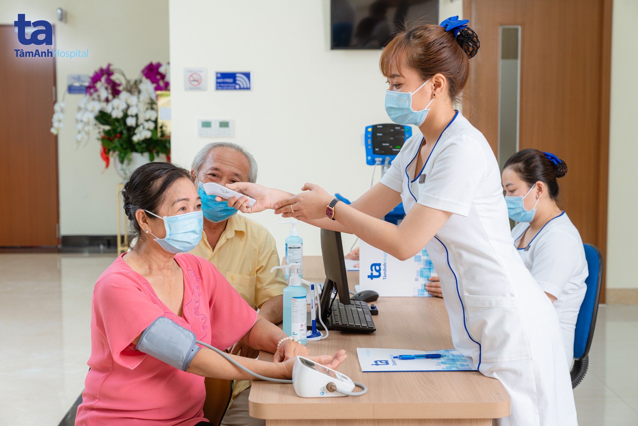 Bác sĩ nữ trong đồng phục trắng đang đo huyết áp cho bệnh nhân nữ lớn tuổi mặc áo hồng, ngồi cạnh người đàn ông lớn tuổi, trong phòng khám có bàn gỗ, máy đo và hoa.