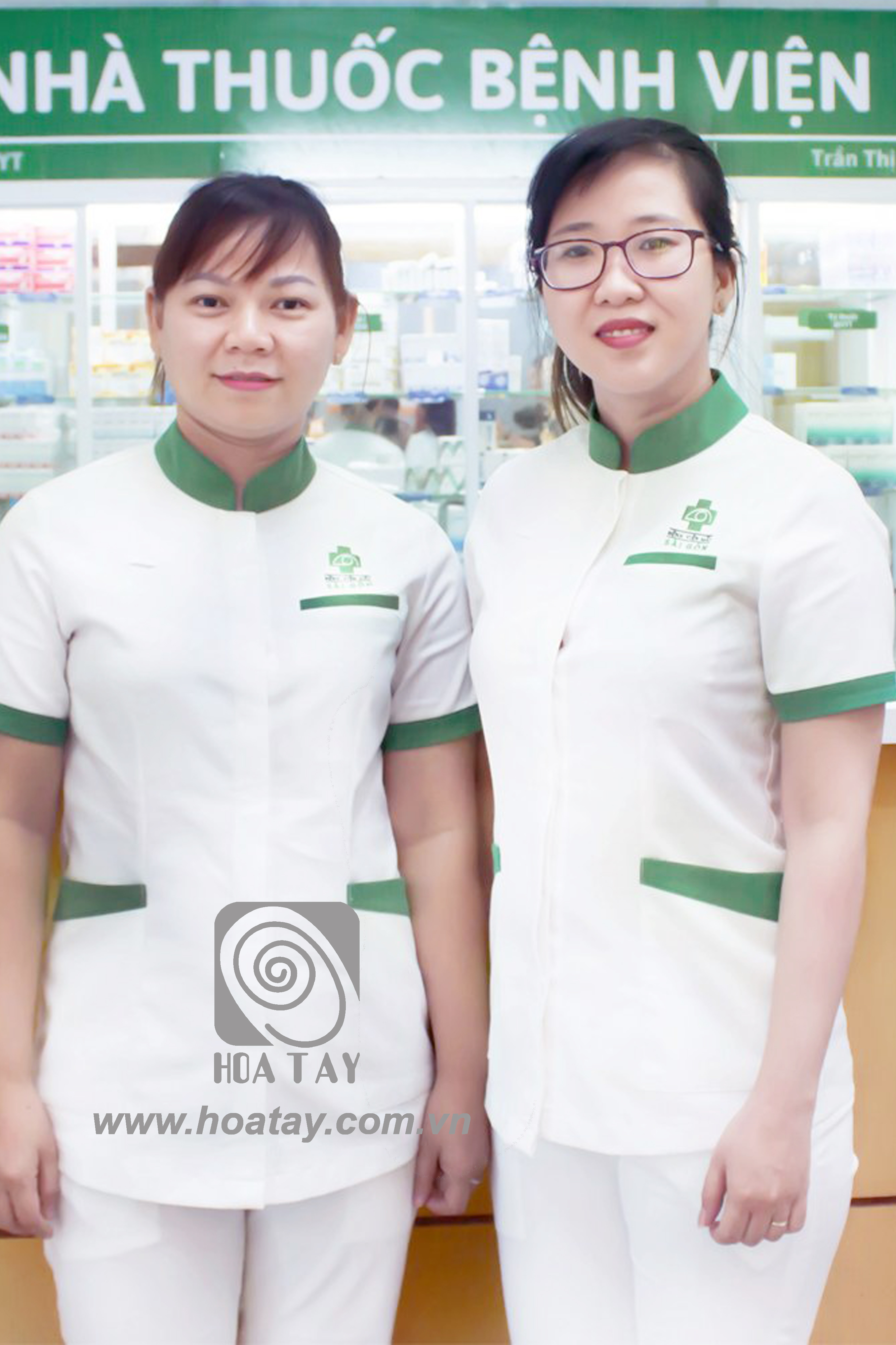 Hai nữ dược sĩ đứng cạnh nhau, mặc áo đồng phục cổ trụ (cổ đứng) màu trắng phối viền xanh lá, tay ngắn, có túi ngực và túi hông, logo thêu trên ngực, bối cảnh quầy thuốc bệnh viện với ánh sáng trắng.