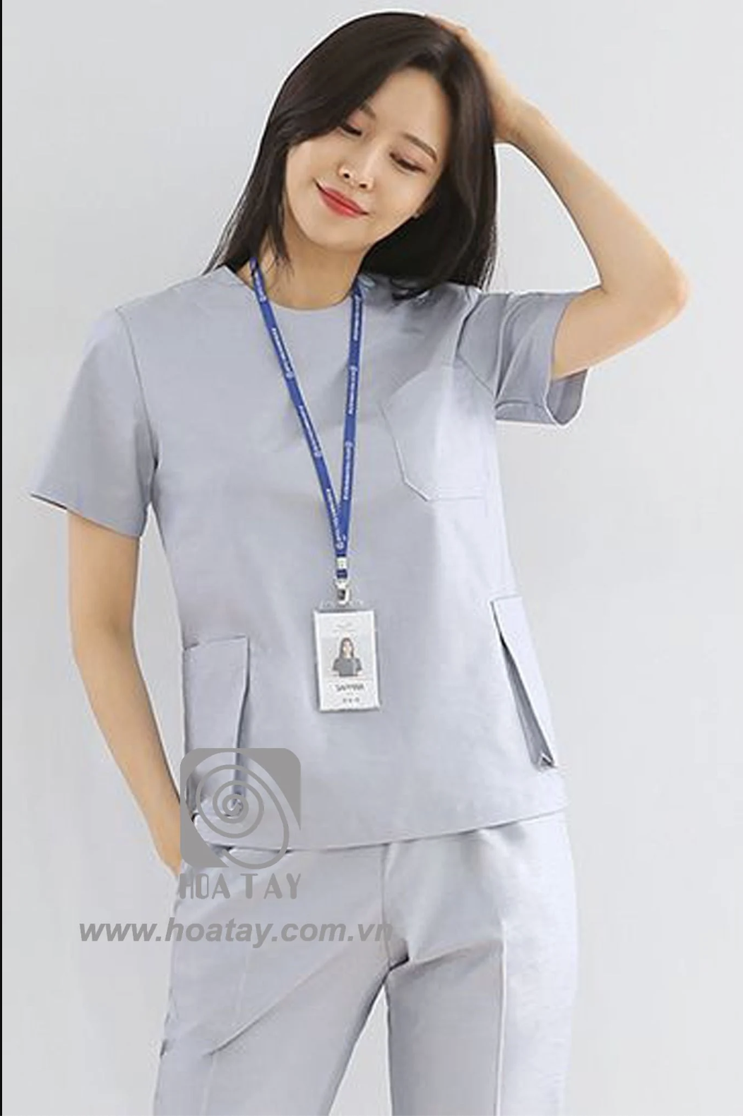Một nữ nhân viên y tế đang đứng, mặc bộ scrub (đồ phẫu thuật) màu xám nhạt, cổ tròn, tay ngắn, có túi ngực và túi hông lớn, đeo thẻ tên, phông nền trắng sáng.