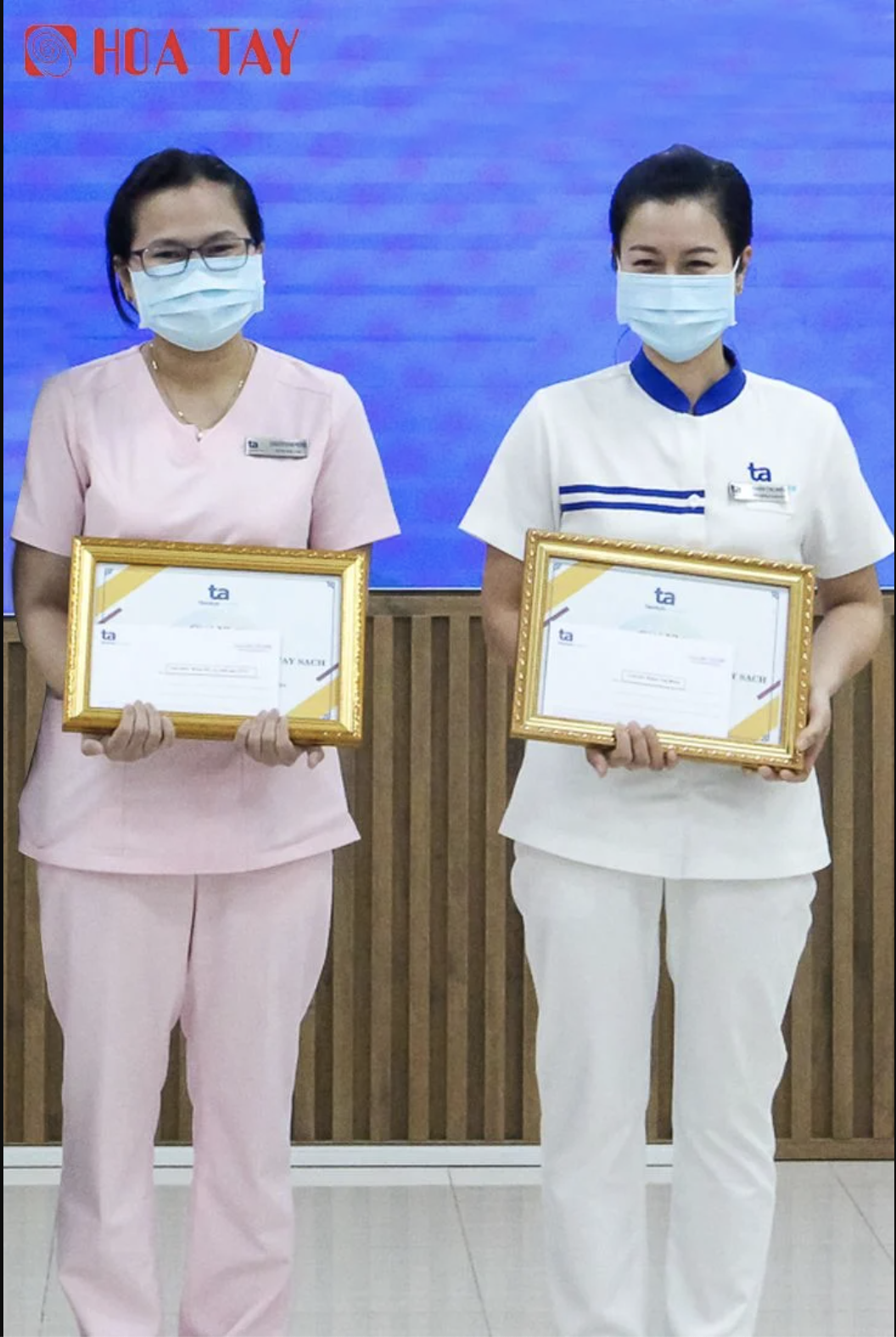 Hai nữ nhân viên y tế (hai người) đang đứng nhận bằng khen, mặc đồng phục cổ chữ V và cổ tròn, màu hồng và trắng, đeo khẩu trang y tế, trong hội trường có phông nền xanh và vách gỗ, ánh sáng đèn huỳnh