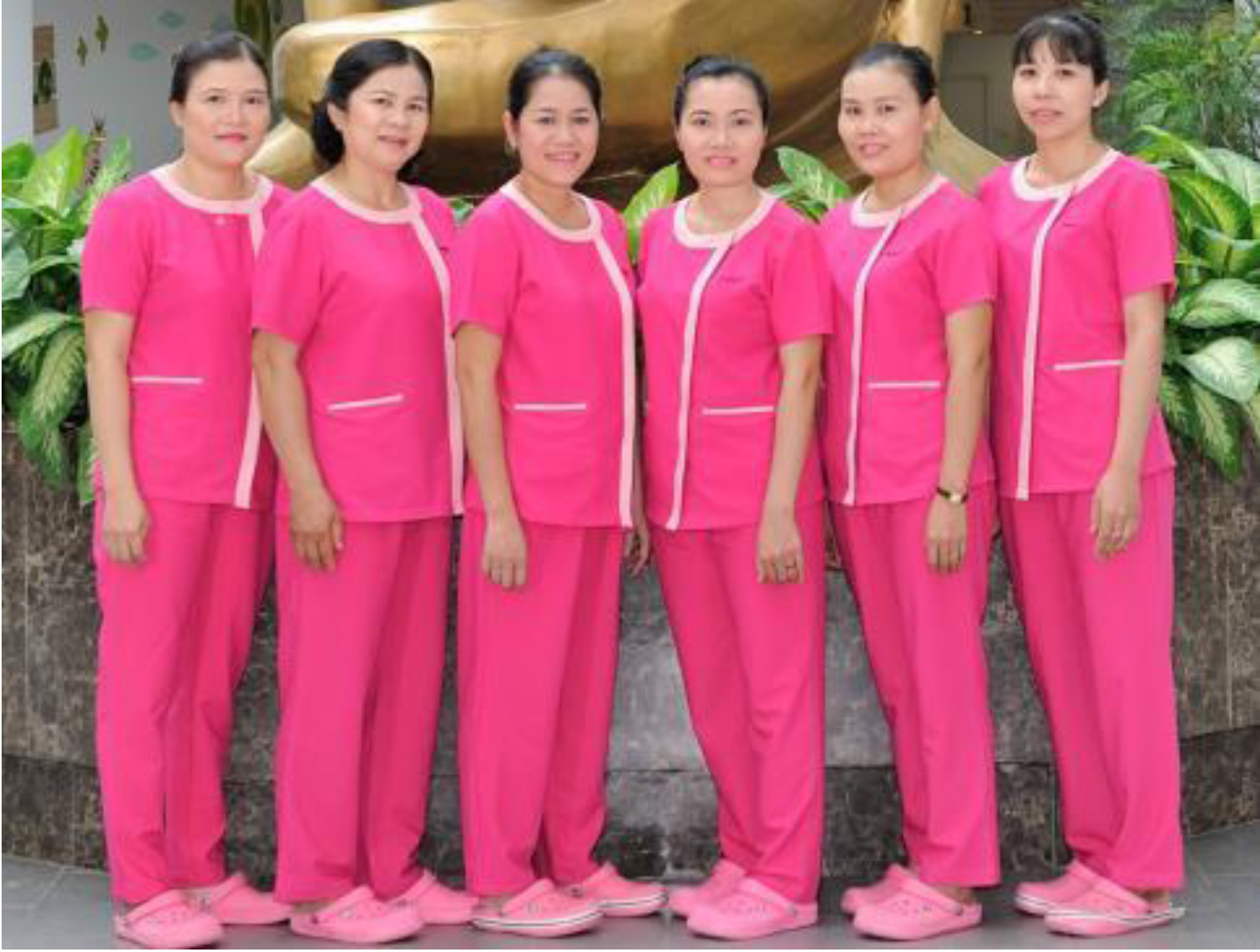 6 nữ nhân viên -> đứng thành hàng -> bộ đồng phục màu hồng với viền trắng, áo ngắn tay, quần dài, giày mềm -> trước tượng vàng và cây xanh