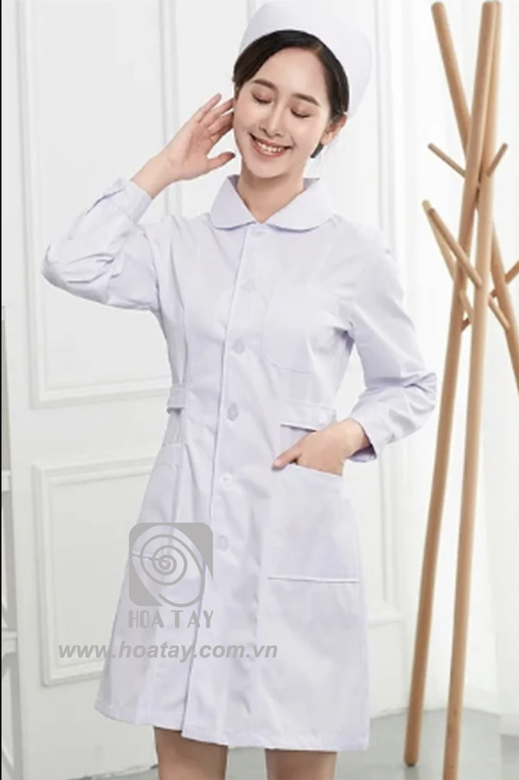 Một nữ y tá (điều dưỡng) đang mỉm cười, mặc áo blouse trắng cổ sen (cổ tròn), tay dài, chất vải cotton trơn, cài khuy giữa, có hai túi hông, đứng trong studio ánh sáng trắng.